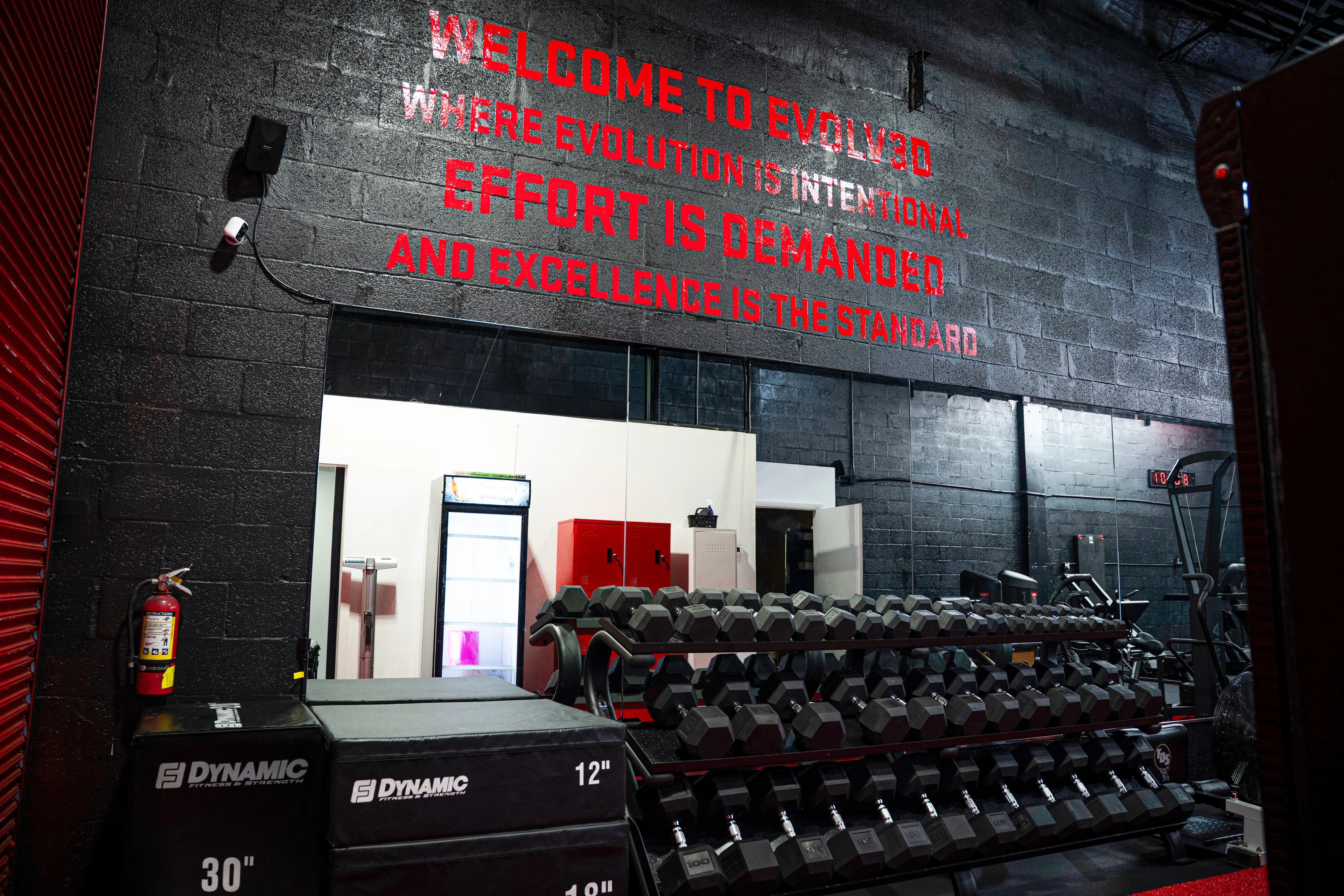 Red lettering on a black brick wall reads: Welcome to Evolv3D, where evolution is intentional, effort is demanded, and excellence is the standard. Dumbbell racks and plyo boxes below.